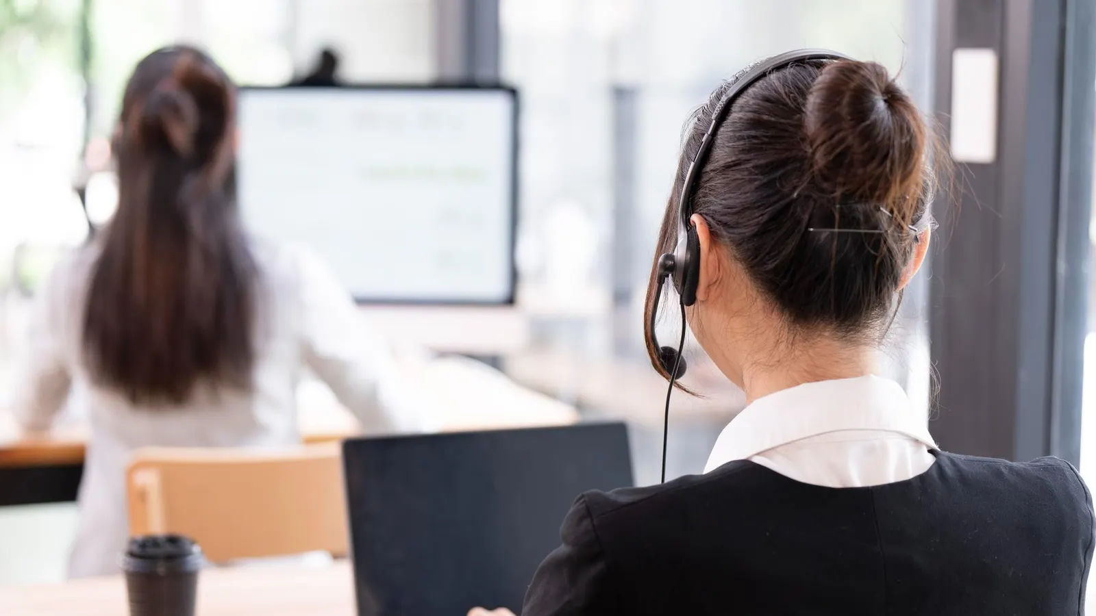 Appointment setter wearing a headset working at a laptop in a bright office, seen from behind