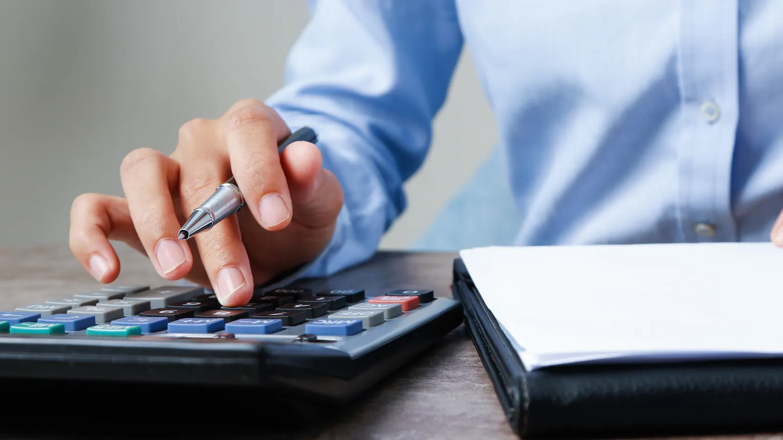 Close-up of hands using a calculator and pen next to a folder of financial documents